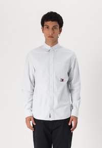 Camisa de botones rayas en blanco y azul claro, fabricada en algodón, con un bolsillo en el pecho y un parche de logo en el lado izquierdo.