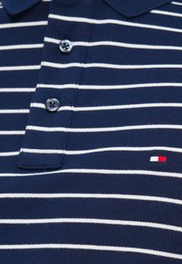 Πόλο μπλούζα σε ναυτικό μπλε με λευκές οριζόντιες ρίγες, τρεις κουμπότρυπες και μικρό κόκκινο, λευκό και μπλε λογότυπο στην περιοχή του στήθους.