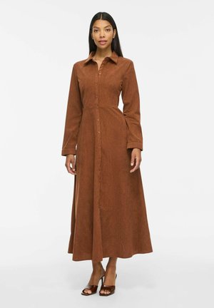 Femme debout portant une robe marron à manches longues boutonnée sur le devant, associée à des chaussures marron à talons ouverts, sur un fond blanc uni.