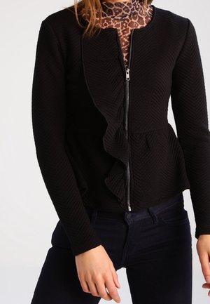 Femme portant une veste zippée noire texturée avec bordures volantées par-dessus un col roulé imprimé léopard marron et un pantalon foncé.