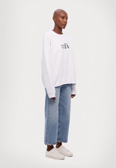 Sweatshirt blanc surdimensionné arborant le mot "ICON" en lettres noires en gras, associé à un jeans droit bleu clair et des baskets blanches. Texture lisse.
