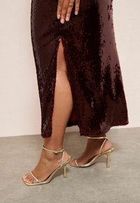 Burgundská šaty s flitry a bočním rozparkem, spárované se zlatými sandály na vysokém podpatku s jemným páskem kolem kotníku.