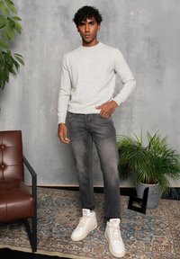 Suéter gris claro de cuello redondo, jeans gris oscuro y zapatillas blancas de caña alta. Ambiente interior con una silla marrón y una planta en maceta.