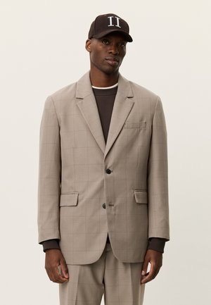 Beige ternet blazer med to knapper, hakkede revers og forlommer, båret over en mørkere top og kombineret med en mørk kasket.