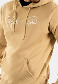 Sudadera marrón con texto bordado blanco "Project x Paris." Cuenta con un bolsillo delantero y cordones ajustables. Material suave y diseño casual.