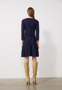 Robe tricotée marine avec des manches trois-quarts et une jupe plissée, associée à des bottes hautes en daim beige et une texture lisse.