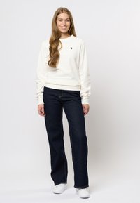 Glunderende vrouw met lang haar, in een witte sweatshirt, donkerblauwe jeans en witte sneakers, staande tegen een effen achtergrond.