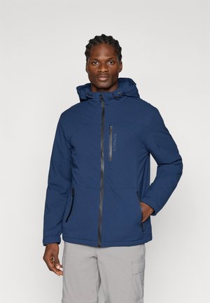 Hombre con cabello trenzado que lleva una chaqueta azul con capucha y bolsillos con cremallera en el pecho y los lados, de pie con una mano en el bolsillo.