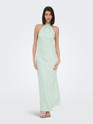 Jeune femme aux cheveux noirs et lisses portant une longue robe sans manches vert pâle à col halter et des sandales beige, debout devant un fond uni.