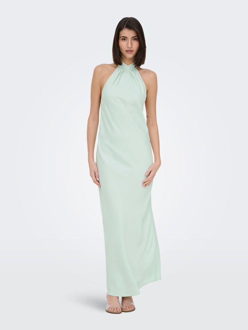 Jeune femme aux cheveux noirs et lisses portant une longue robe sans manches vert pâle à col halter et des sandales beige, debout devant un fond uni.