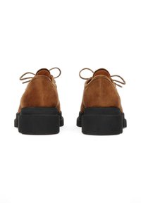 Pantofi din suede maro, cu un design cu vârf rotund, tălpi negre groase și fundațe legate la spate pentru prindere. Textură netedă, fără modele.