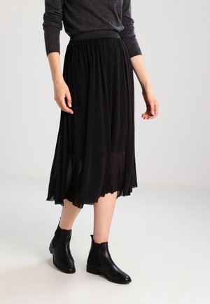 Femme portant un haut gris à manches longues, une jupe midi plissée noire et des bottines noires marchant sur un fond clair.