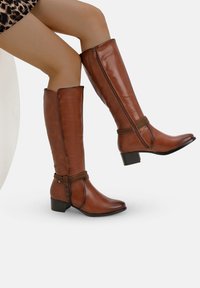 Botas de cuero marrón hasta la rodilla con punta ligeramente afilada, tacón plano y detalle decorativo en la muñeca. Textura suave, acabado brillante.