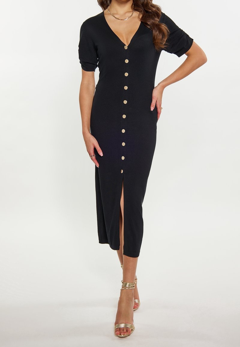 Femme portant une robe midi noire avec des boutons sur le devant et une fente, associée à des sandales à talons hauts dorées et des bijoux.