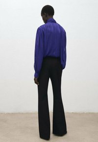 Blusa viola plissettata con maniche lunghe e colletto alto, abbinata a pantaloni neri svasati, vista da dietro su uno sfondo neutro.