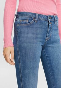 Jeans de denim azul ajustados con costuras naranjas en contraste, bolsillos frontales y una cinturilla de altura media, combinados con un suéter de punto rosa.