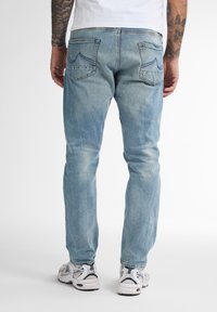 Jeans de mezclilla azul claro con corte recto, detalles desgastados y dobles bolsillos traseros con diseños cosidos. Llevados con zapatillas blancas.