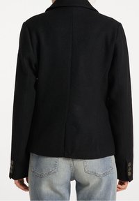 Czarna wełniana kurtka z usztywnianym kołnierzem, dopasowanym krojem i mankietami zapinanymi na guziki. Gładka faktura bez widocznych wzorów. Noszona na jasnym denimu.