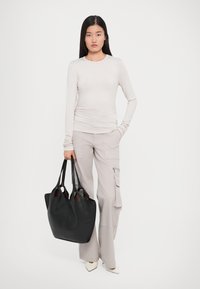 Femme portant un haut beige à manches longues et un pantalon cargo large, tenant un grand sac fourre-tout en cuir noir, se tenant contre un fond blanc.