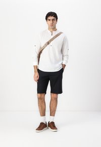 Bílá dlouhá halenka s knoflíky, černé šortky, hnědé tenisky a béžová kabelka přes rameno na jednobarevném pozadí.