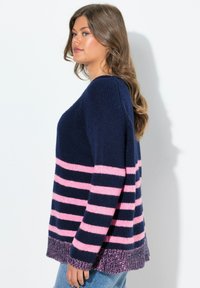 Vrouw met lang bruin haar, gekleed in een marineblauwe trui met roze horizontale strepen en lichtblauwe jeans, staande tegen een witte achtergrond.