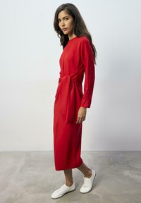 Rode jurk met lange mouwen en een geknoopte detail in de taille, gemaakt van zachte stof, gecombineerd met witte sneakers, staande op een betonnen vloer.