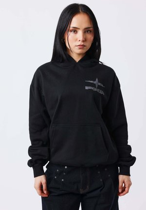 Jonge vrouw met lang zwart haar, gekleed in een zwarte hoodie met een subtiel sterrenpatroon en donkere broek met kleine cirkelvormige details.