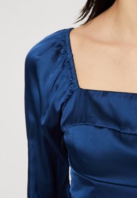 Námořnická modrá saténová šaty s čtvercovým výstřihem, nabíranými detaily na ramenou a dlouhými rukávy. Hladká textura a přiléhavý silueta.