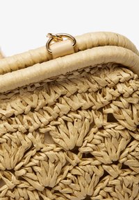 Tkaná slaměná kabelka s texturovaným designem v světle béžové barvě. Zakulacený vrch s akcenty zlaté kovové barvy a hladkým koženým okrajem.