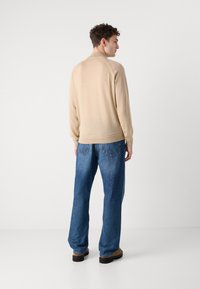 Beige lett strikkegenser med ribbestrikket nederkant og mansjetter, sammen med løse blå denimjeans og brune sko, sett fra baksiden.