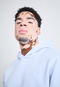Šviesiai mėlyna pūkinė striukė, pagaminta iš minkštos medžiagos, su balta Jumpman logotipu ant krūtinės. Artimas veido kadras su išskirtiniu odos raštu.