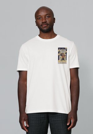 Witte katoenen t-shirt met een afbeelding van een koninklijke figuur op de linkerborst, met gedurfde kleuren en gedetailleerde illustratie. Korte mouwen en ronde hals.