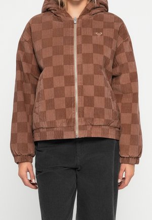 Osoba nosząca brązową kraciastą kurtkę z kapturem z aksamitu sztruksowego z zamkiem i elastycznymi mankietami, w połączeniu z czarnymi spodniami.