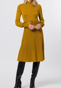Senapsgul stickad klänning med hög hals, bältad midja och vertikala sömdetaljer; tillsammans med svarta över knästövlar.