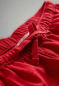 Rote Stoffshorts mit einem gerafften Bund und einer gebundenen Schleifendetail. Die Textur ist weich, mit sichtbaren Nähten entlang der Kanten.
