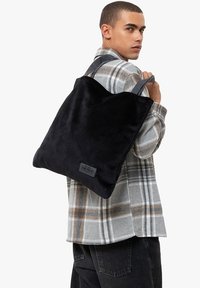 Svart mocka tote-väska med grå handtag, fyrkantig form; har en liten logotypapplikation. Modellen bär en rutmönstrad skjorta med svarta byxor.