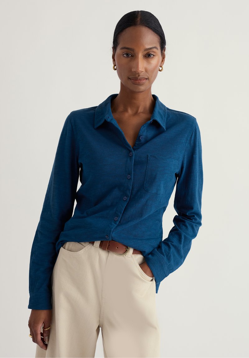 Camisa azul de manga larga con botones, cuello y un bolsillo en el pecho, combinada con pantalones crema y un cinturón marrón.