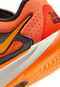 Athletic shoe featuring vibrant orange, black, and white materials. Textured accents and perforations visible on the synthetic upper.