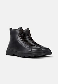 Une paire de bottes en cuir noir à lacets avec des semelles épaisses à crampons sur un fond gris clair uni.