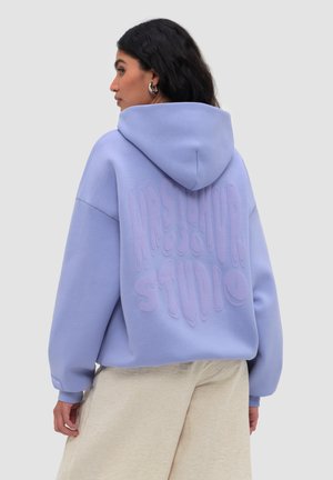 Kobieta o ciemnych włosach ubrana w oversize lawendową bluzę z kapturem z wypukłym napisem "Mastermind Studio" na plecach, beżowe spodnie oraz srebrne kolczyki-koła.