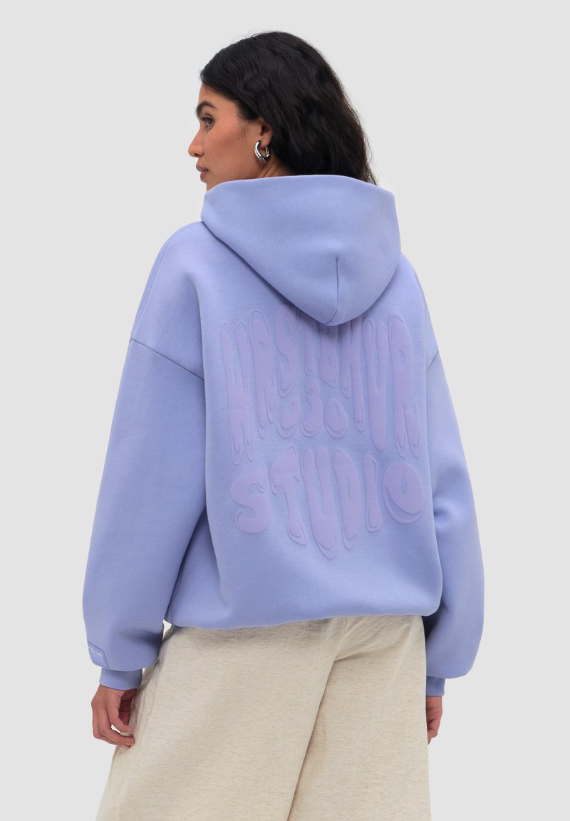 Femme aux cheveux foncés portant un sweat à capuche lavande oversized avec texte en relief "Mastermind Studio" dans le dos, pantalon beige et boucles d'oreilles créoles argentées.