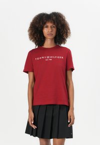 Raudona medvilninė marškinėliai su trumpomis rankovėmis, puoštas baltu tekstu "TOMMY HILFIGER EST. 1985" per krūtinę, derinami su juoda klostuota sijonu.