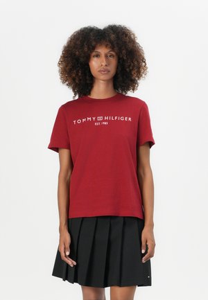 T-shirt en coton rouge à manches courtes avec le texte blanc "TOMMY HILFIGER EST. 1985" sur la poitrine, associé à une jupe plissée noire.