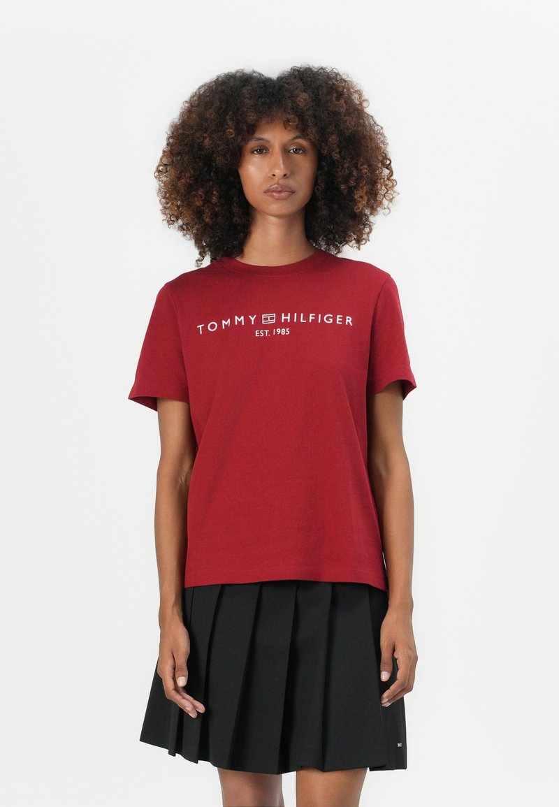 Raudona medvilninė marškinėliai su trumpomis rankovėmis, puoštas baltu tekstu "TOMMY HILFIGER EST. 1985" per krūtinę, derinami su juoda klostuota sijonu.