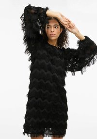 Robe noire avec des motifs ondulés texturés, présentant des manches à franges et une coupe décontractée. Le tissu transparent ajoute de la dimension.