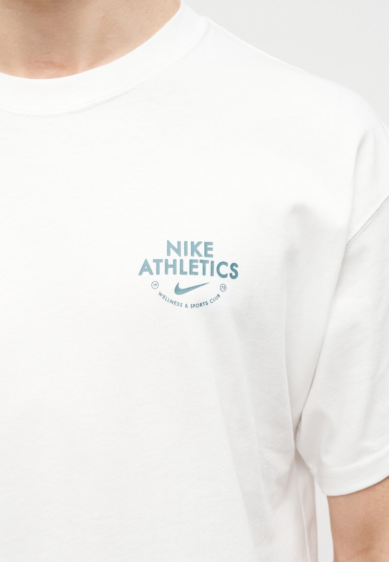 Maglietta bianca con la scritta "Nike Athletics", logo swoosh e "Wellness & Sports Club" sull'area del petto sinistro indossata da una persona.