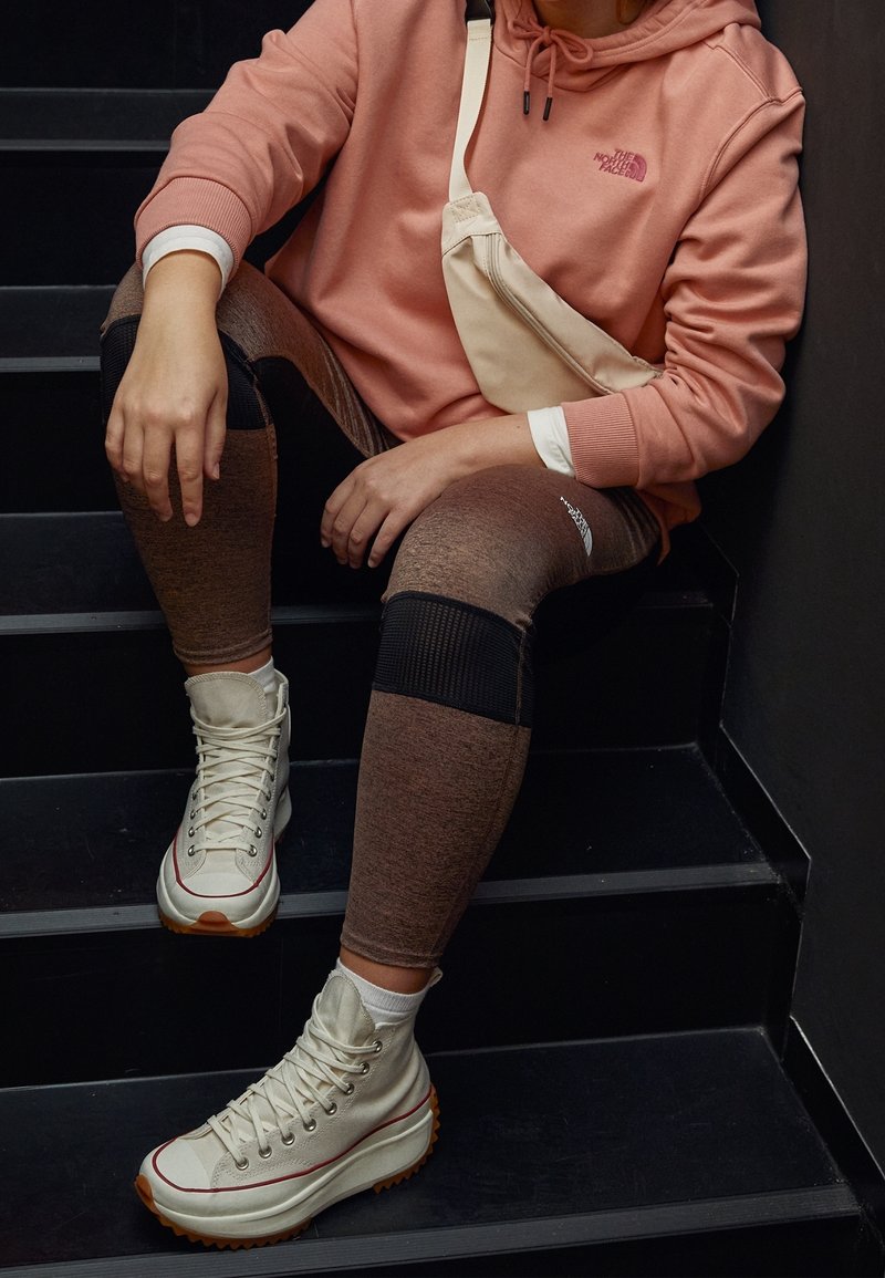 Personne assise sur des escaliers noirs, portant un sweat à capuche rose de la marque North Face, des leggings marron avec des patchs noirs, des baskets montantes blanches et un sac bandoulière beige.