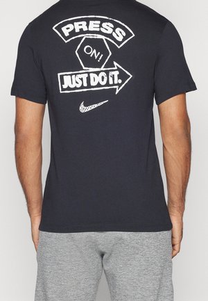 Mann trägt ein schwarzes T-Shirt mit weißem Text "PRESS ON! JUST DO IT." und Nike-Swoosh auf dem Rücken, kombiniert mit grauen Shorts.