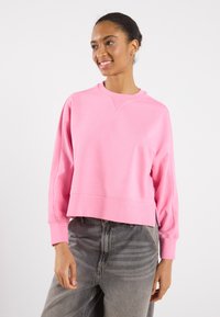 Felpa rosa tagliata con maniche lunghe, polsini a costine e scollatura a girocollo. Tessuto morbido e design minimalista. Abbinata a pantaloni grigi chiari.