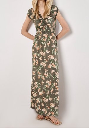 Femme portant une robe longue vert olive avec imprimé floral rose et manches courtes bouffantes, debout, mains sur les hanches et sandales beiges.